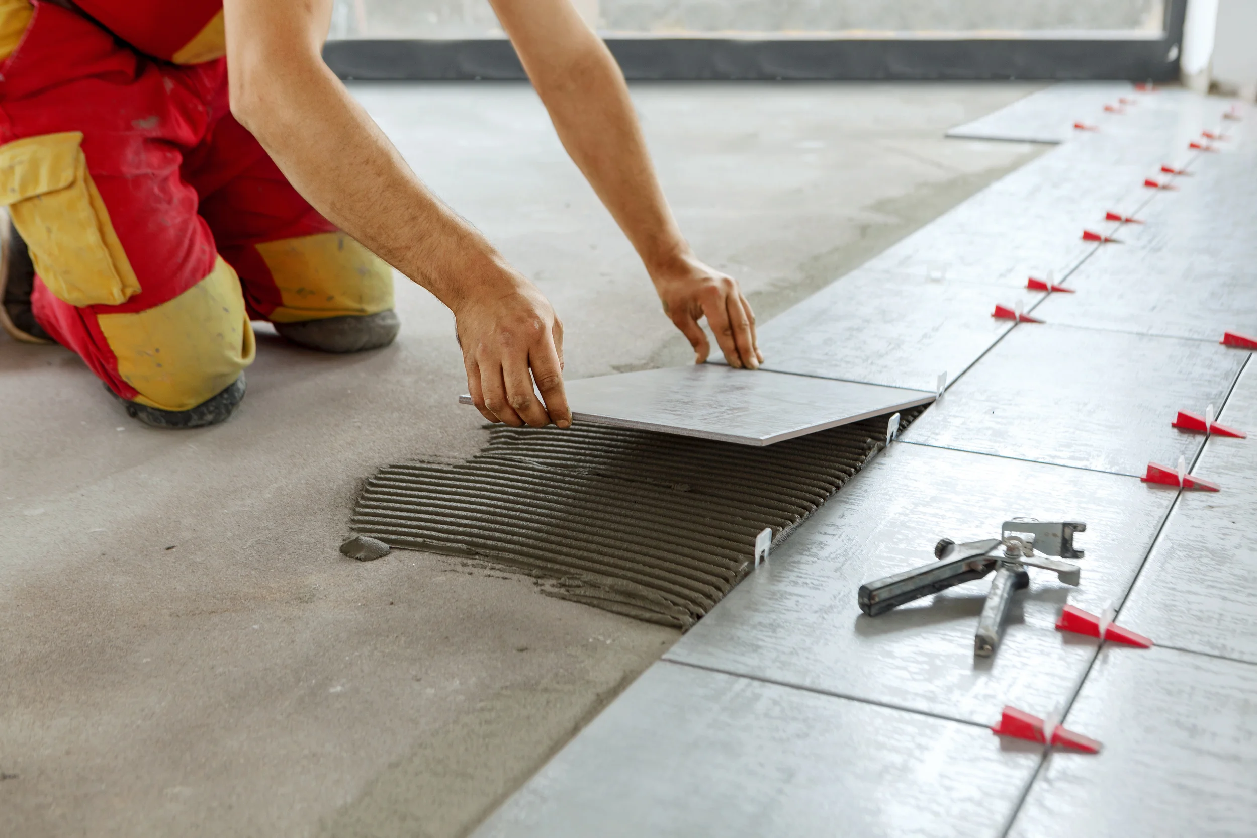 Worker laying a large floor tile onto combed mortar with red tile spacers